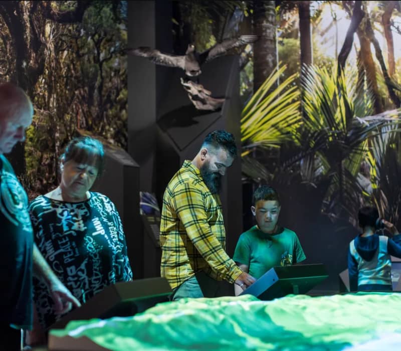 People engaging with interactive displays in a museum exhibit with a forest background and bird model.