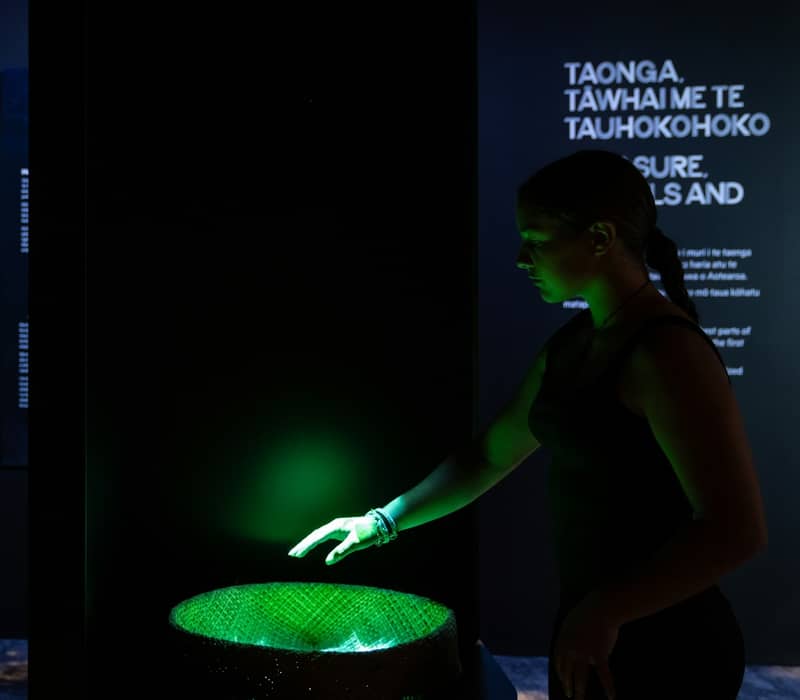 Two women interacting with illuminated cultural exhibits in a dark museum room.