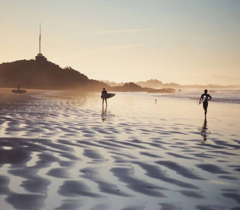 Surfers walking on a beach at sunset carrying surfboards with rippled sand and waves in the background.