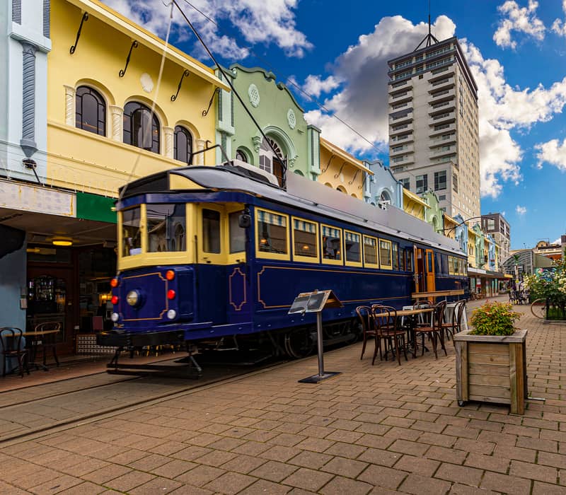Christchurch Blue Tram on a brick road with colourful buildings behind, benches and plants in the foreground with a blue sky 