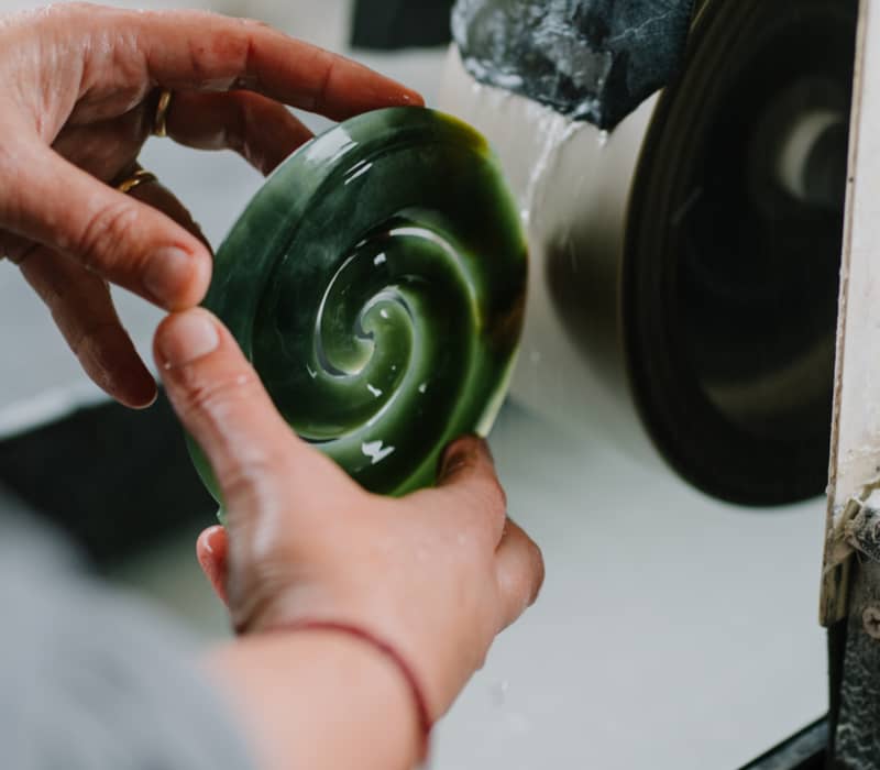 Hands polishing a green jade piece with a swirl pattern using a wet grinding machine.