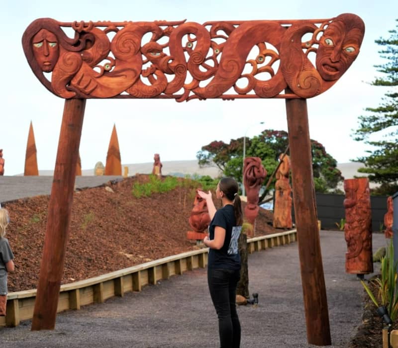 A group of visitors, including children, listening to a Māori guide in front of a monumental, intricately carved wooden gateway at Manea Footprints of Kupe.