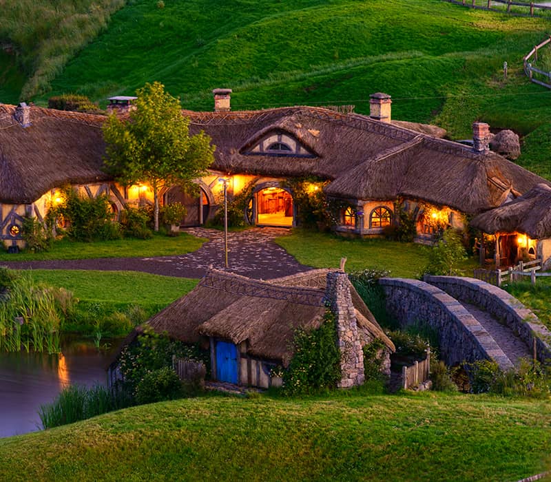 Hobbiton Movie Set cottages and stone bridge lit warmly at dusk surrounded by green hills and river.