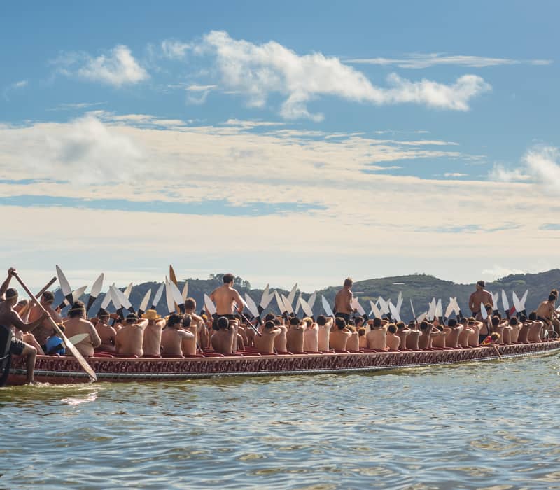 People paddling a traditional waka canoe on water with hills and blue sky in background.