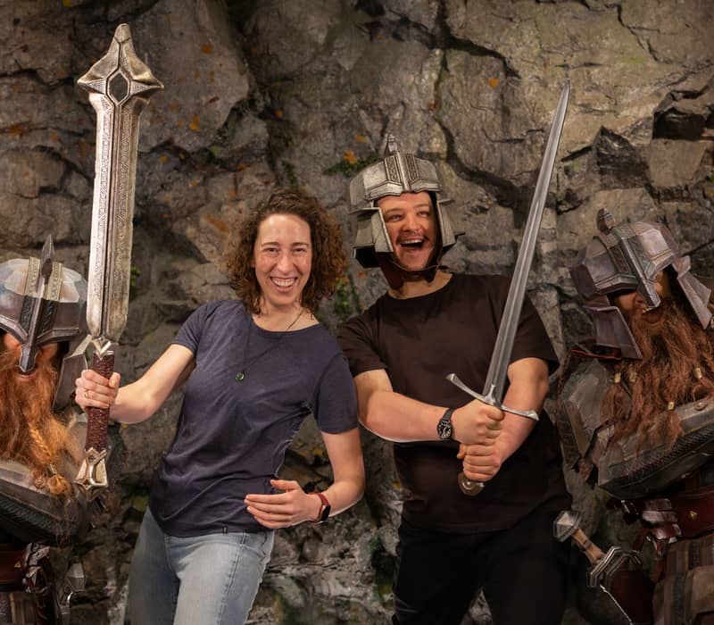 Two people smiling and holding swords while wearing metal helmets, posing with dwarves in armor against a rocky background.