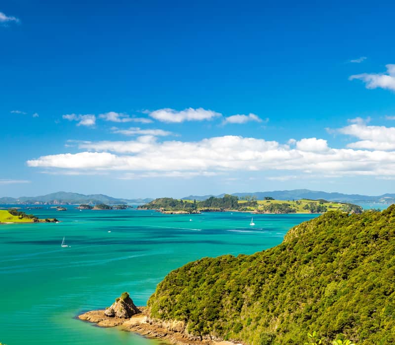 Coastal landscape with green hills, turquoise sea, sailboats, and blue sky with clouds.