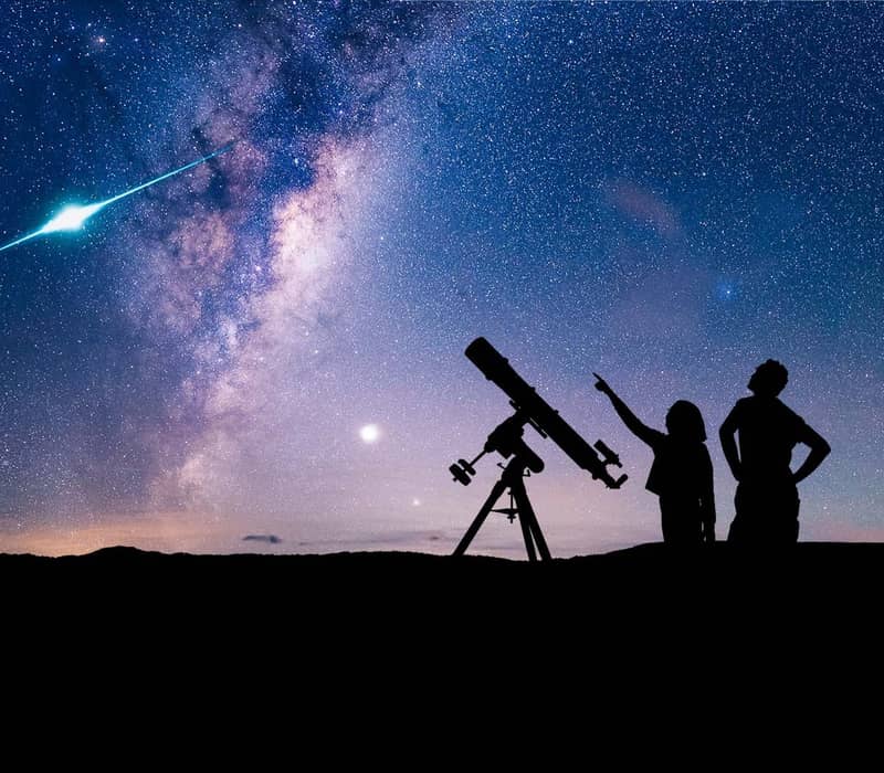 Silhouettes of two people and a telescope under a starry night sky with the Milky Way and a bright meteor.