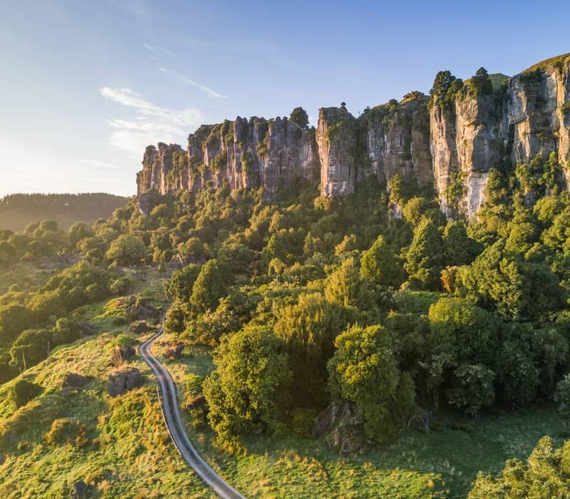 Sunlit cliffs with green forest and a winding trail in a scenic natural landscape.