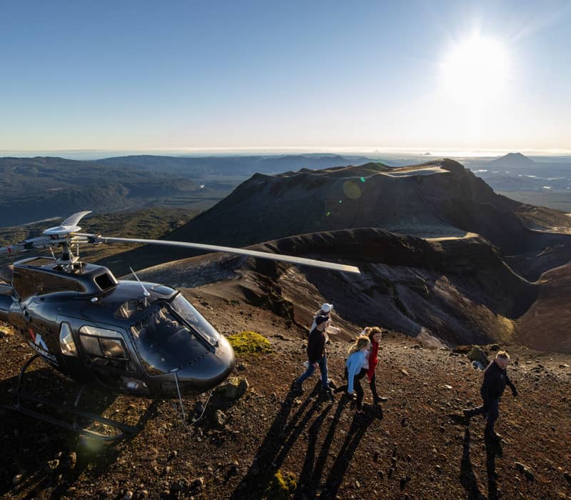 Tourists walking near a helicopter on a volcanic crater under bright sunshine.