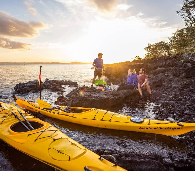 Two yellow kayaks on water near rocky shore with couple enjoying a picnic at sunset.