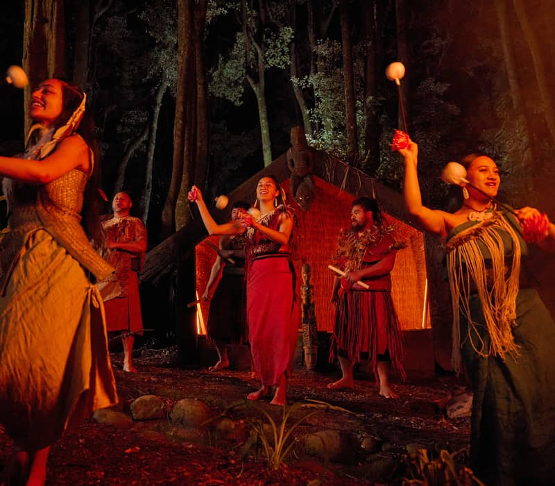 Māori performers in traditional dress perform a poi dance on a forest stage at night, with a carved wharenui and towering trees illuminated in the background.