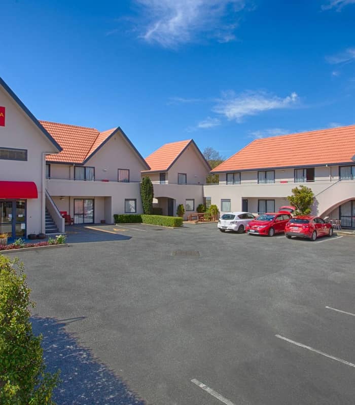 Exterior of Bella Vista Motel with red roofs, cars parked and clear blue sky with clouds.