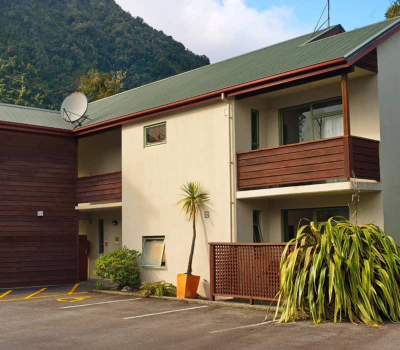 Two-story motel building with balconies and parking in front, with a green mountain behind.