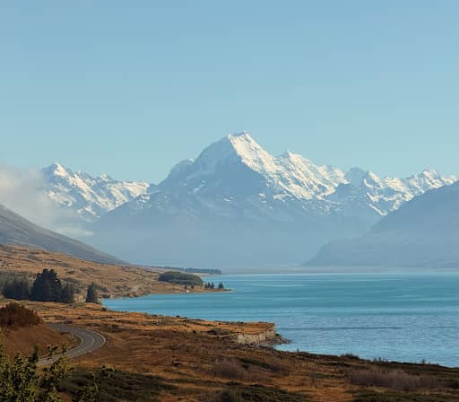 A vertical shot of a winding lakeside road leading toward the towering, snow-capped peak of Aoraki / Mount Cook across the bright blue waters of Lake Pukaki.