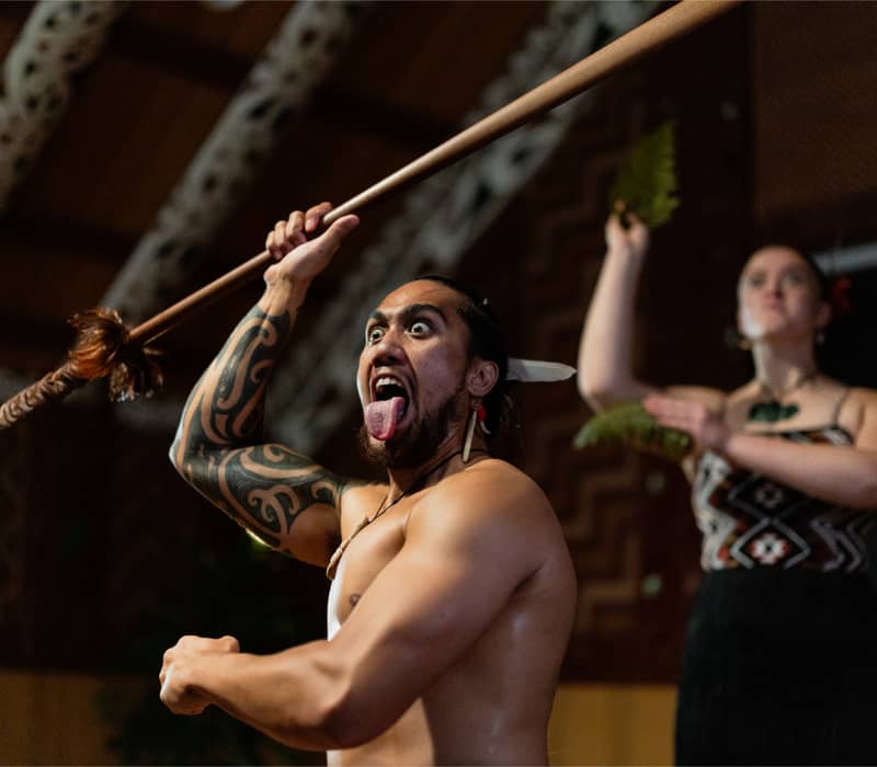 Maori warrior performing a traditional haka with spear and fierce expression.