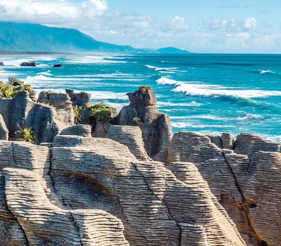 Punakaiki Pancake Rocks on New Zealand's west coast with ocean and mountains.