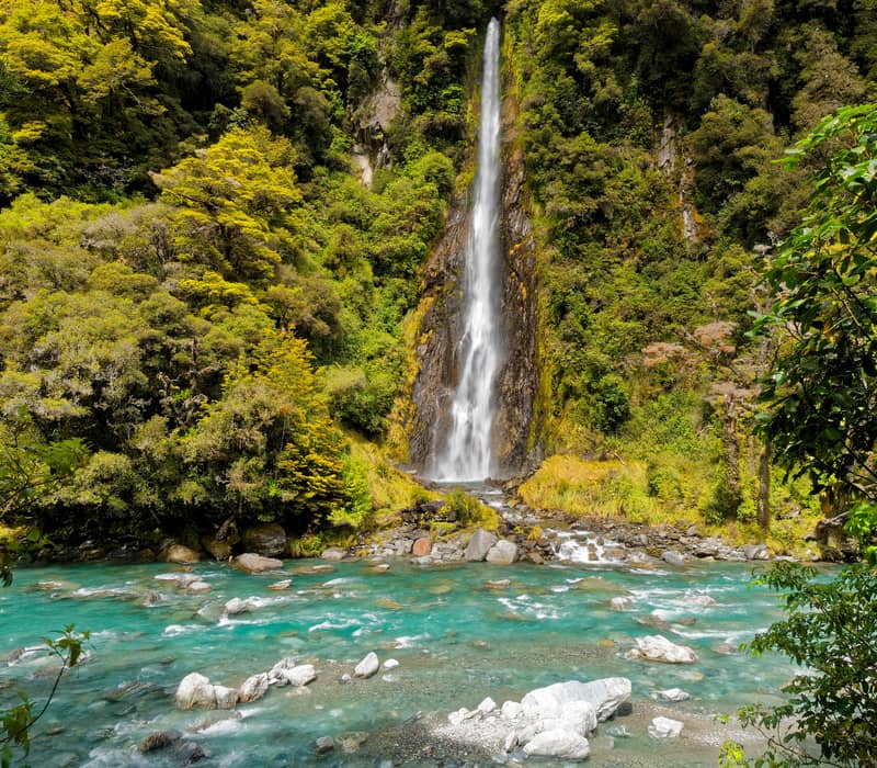 Tall waterfall cascading into bright blue river with green forest surrounding.