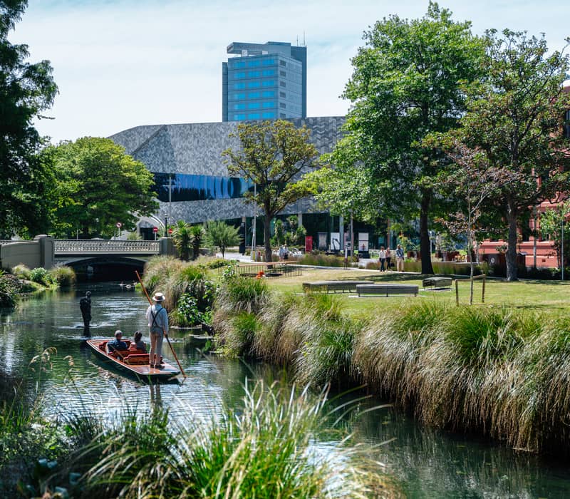People punting on a river in a green park with buildings and a bridge in the background on a sunny day.