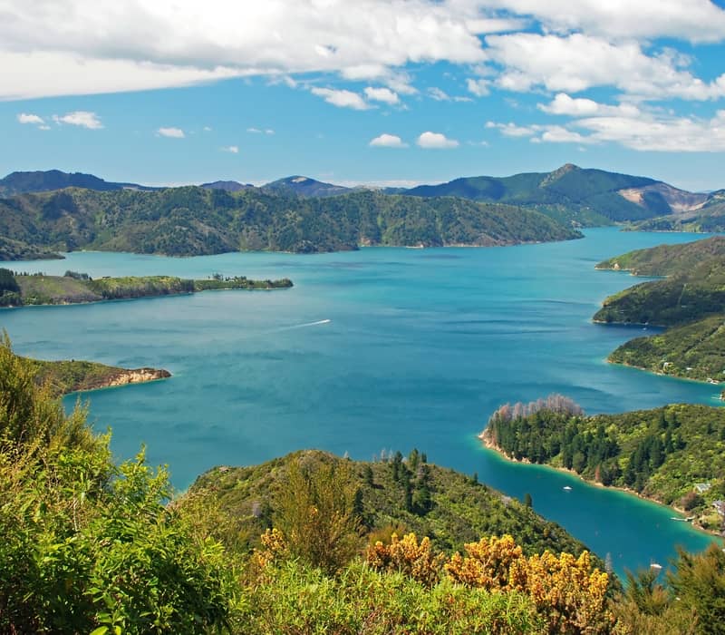Scenic lake with green mountains and forests under a blue sky with clouds.