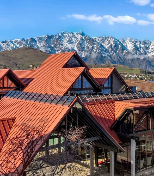 Hotel with red roofs near lakefront and snow-covered mountains in the background under a clear sky.