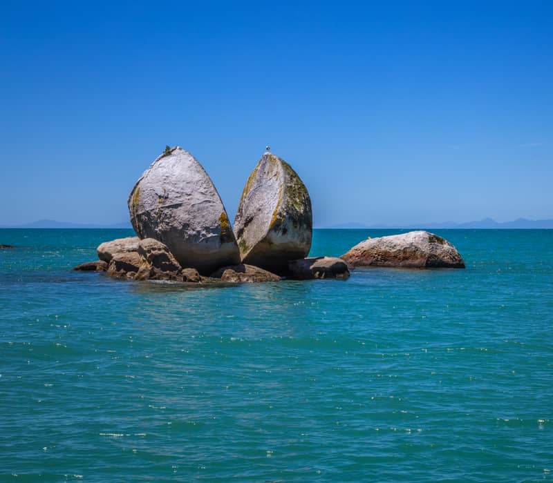 A large split granite boulder in the ocean under a clear blue sky.