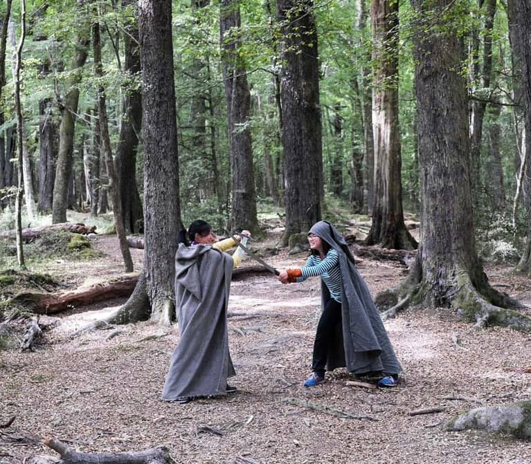 Two people wearing cloaks mock sword fighting in a dense leafy forest.