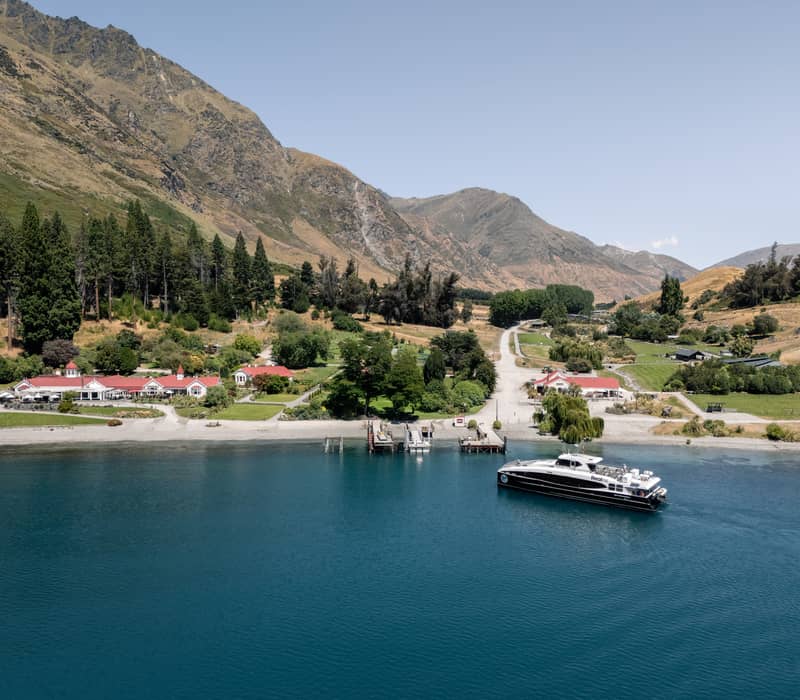 Boat on lake near pier with mountains and green farm buildings beyond under clear sky.