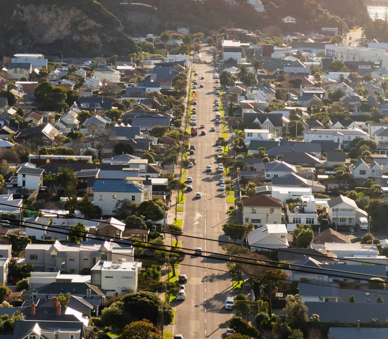 An elevated view looking down a straight suburban street lined with houses toward a green hillside and the coast in New Zealand.