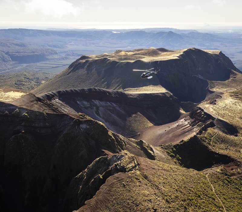 Helicopter flying over the crater of Mount Tarawera volcano with rugged terrain and distant mountains