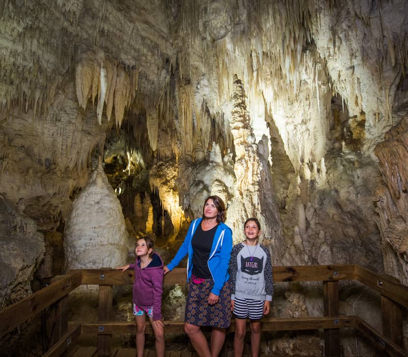 A mother and her two young daughters standing on a wooden viewing platform inside a cave, looking up in awe at thousands of delicate, illuminated stalactites.