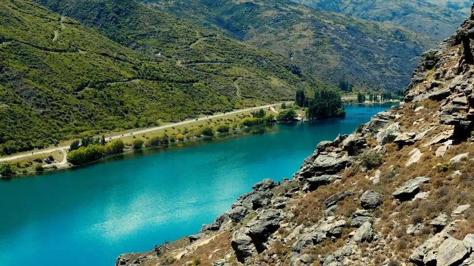 Blue river flowing beside a winding road with green hills and rocky cliffs.
