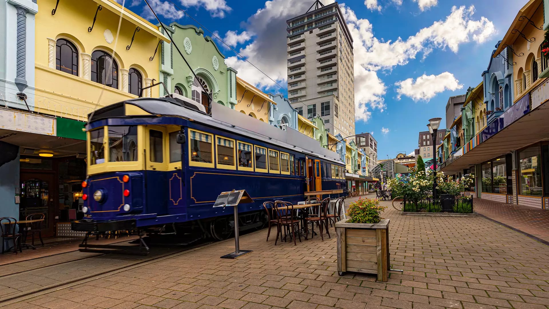 Christchurch Blue Tram on a brick road with colourful buildings behind, benches and plants in the foreground with a blue sky 