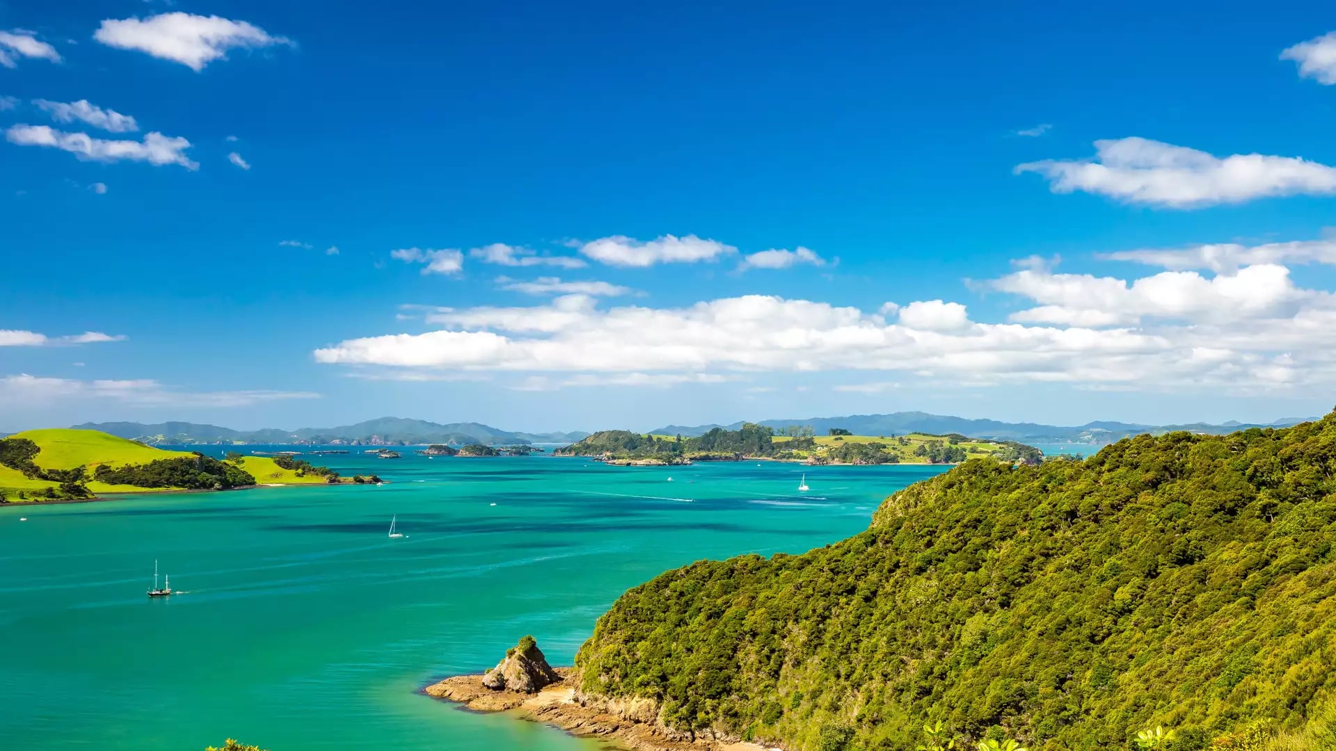 Coastal landscape with green hills, turquoise sea, sailboats, and blue sky with clouds.