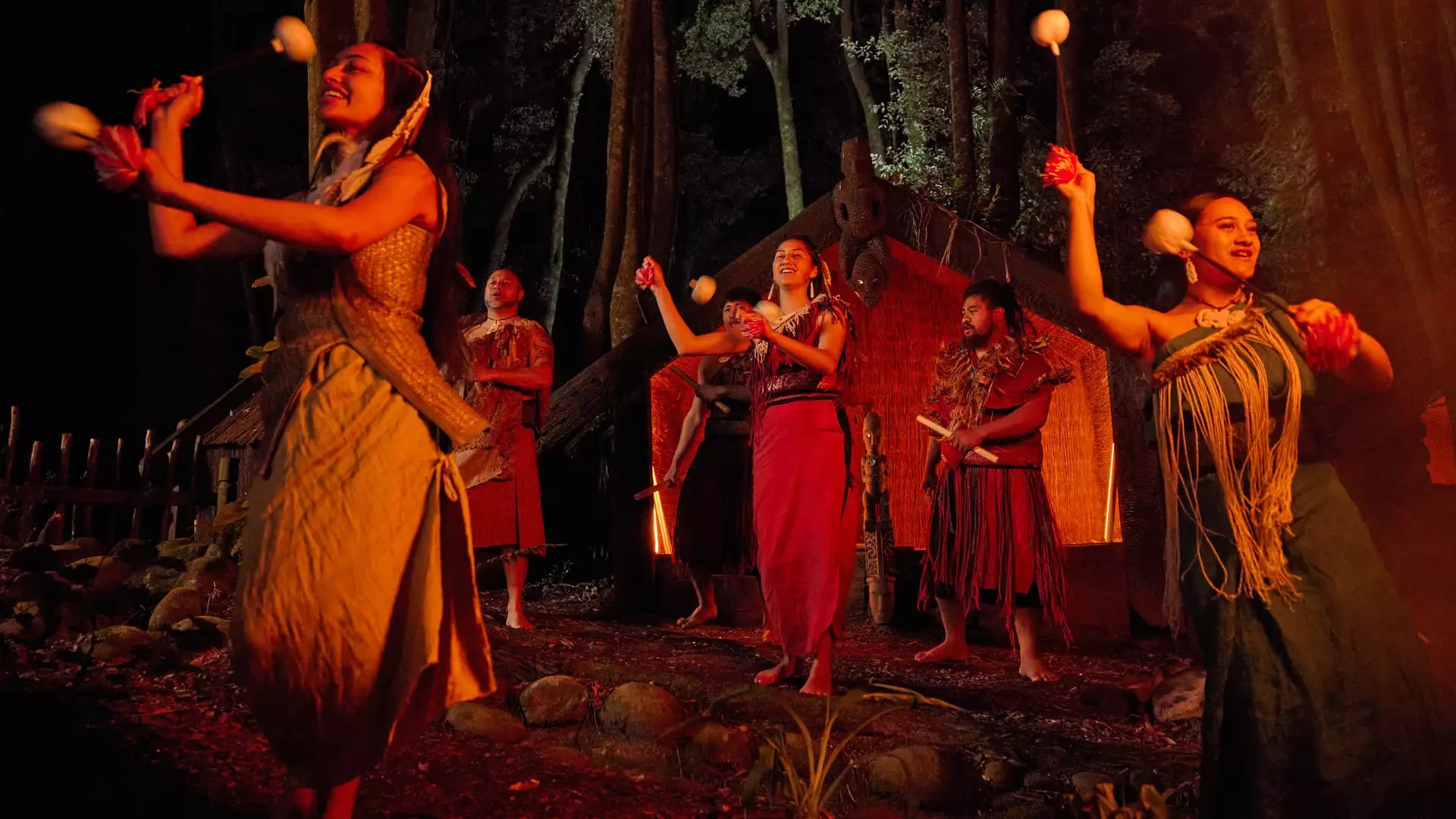 Māori performers in traditional dress perform a poi dance on a forest stage at night, with a carved wharenui and towering trees illuminated in the background.