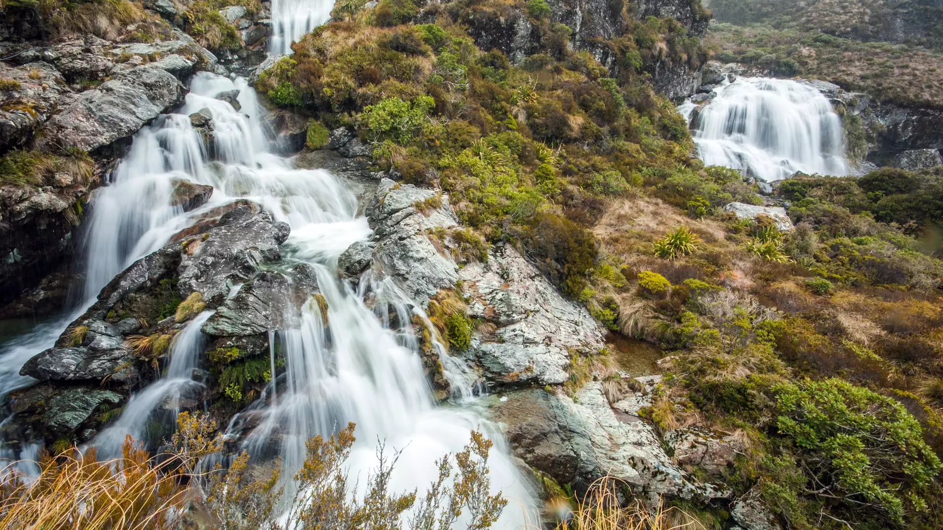 Waterfalls flowing over rocks with green shrubs and moss on a mountain landscape