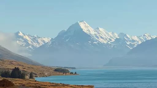 A vertical shot of a winding lakeside road leading toward the towering, snow-capped peak of Aoraki / Mount Cook across the bright blue waters of Lake Pukaki.