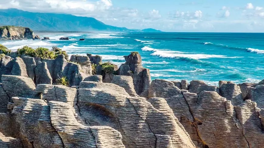 Punakaiki Pancake Rocks on New Zealand's west coast with ocean and mountains.