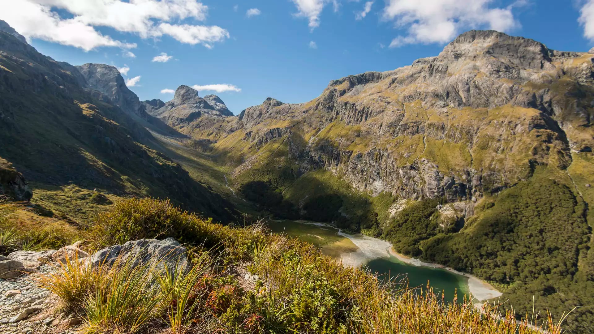 Mountain valley with a lake, green bushes, and blue sky with clouds.