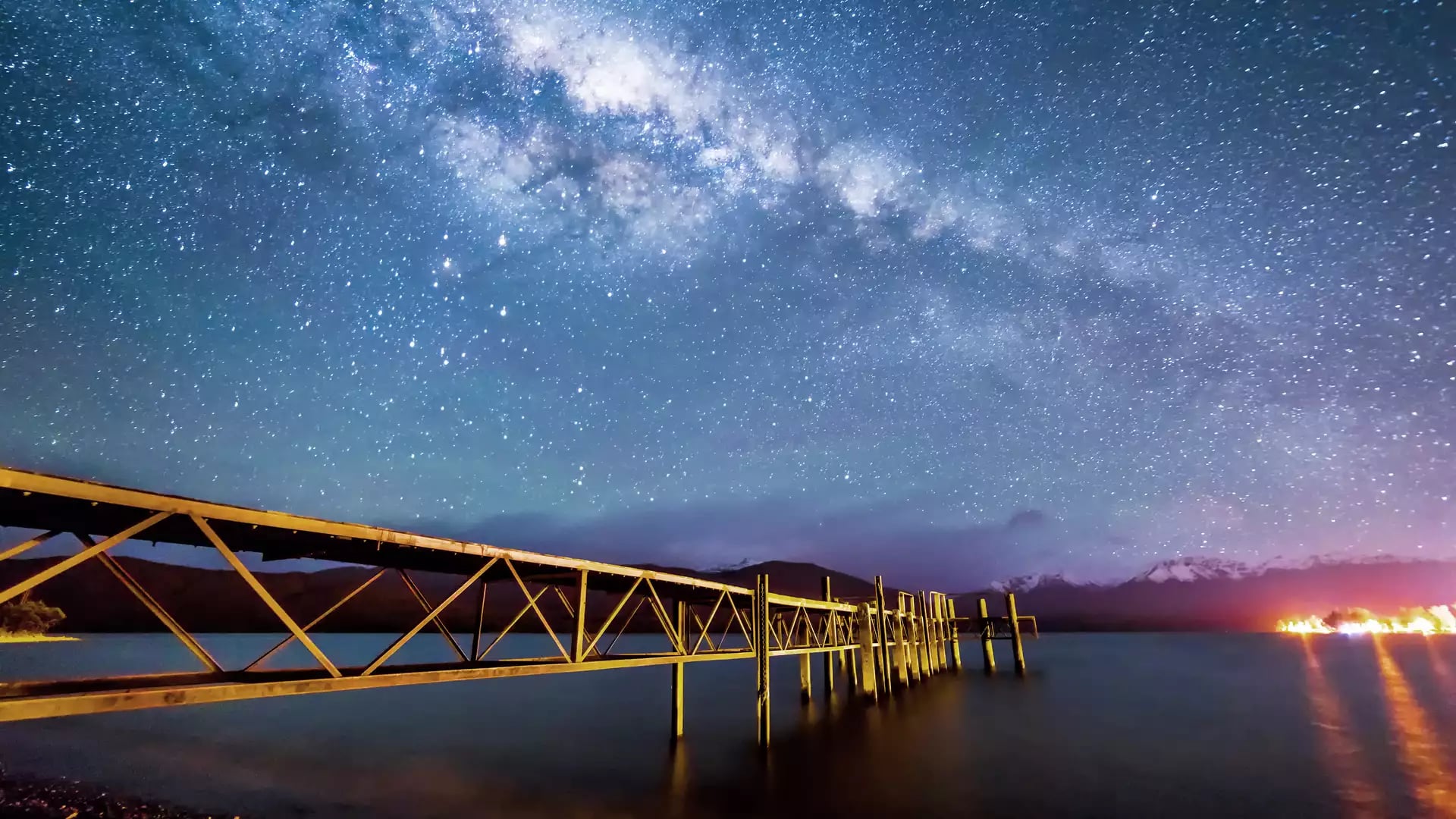 The Milky Way galaxy glows brilliantly above a long wooden jetty stretching into the calm, dark waters of Lake Te Anau.
