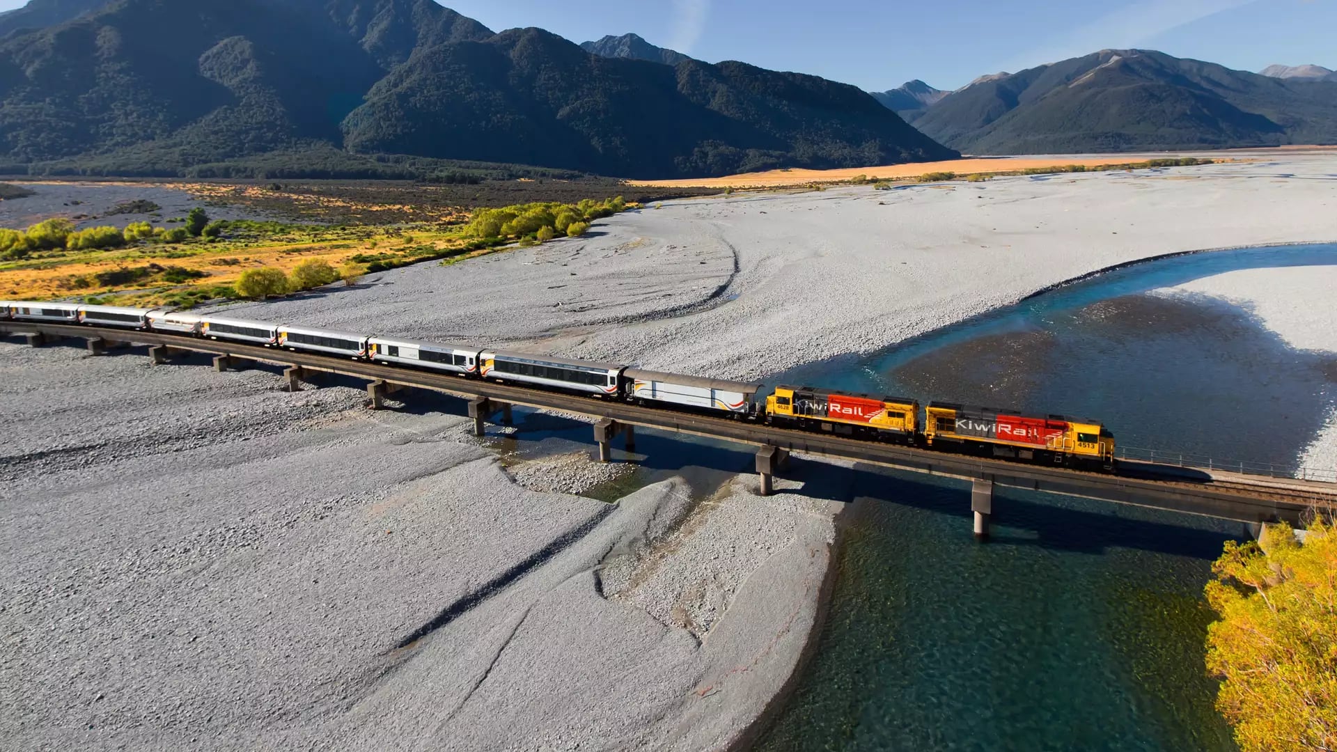 Train crossing bridge over river with mountains in background on a sunny day