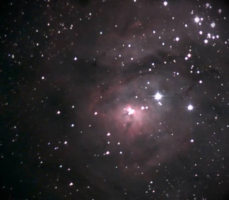 A telescope view of the pink and purple Lagoon Nebula surrounded by bright stars in a dark night sky.
