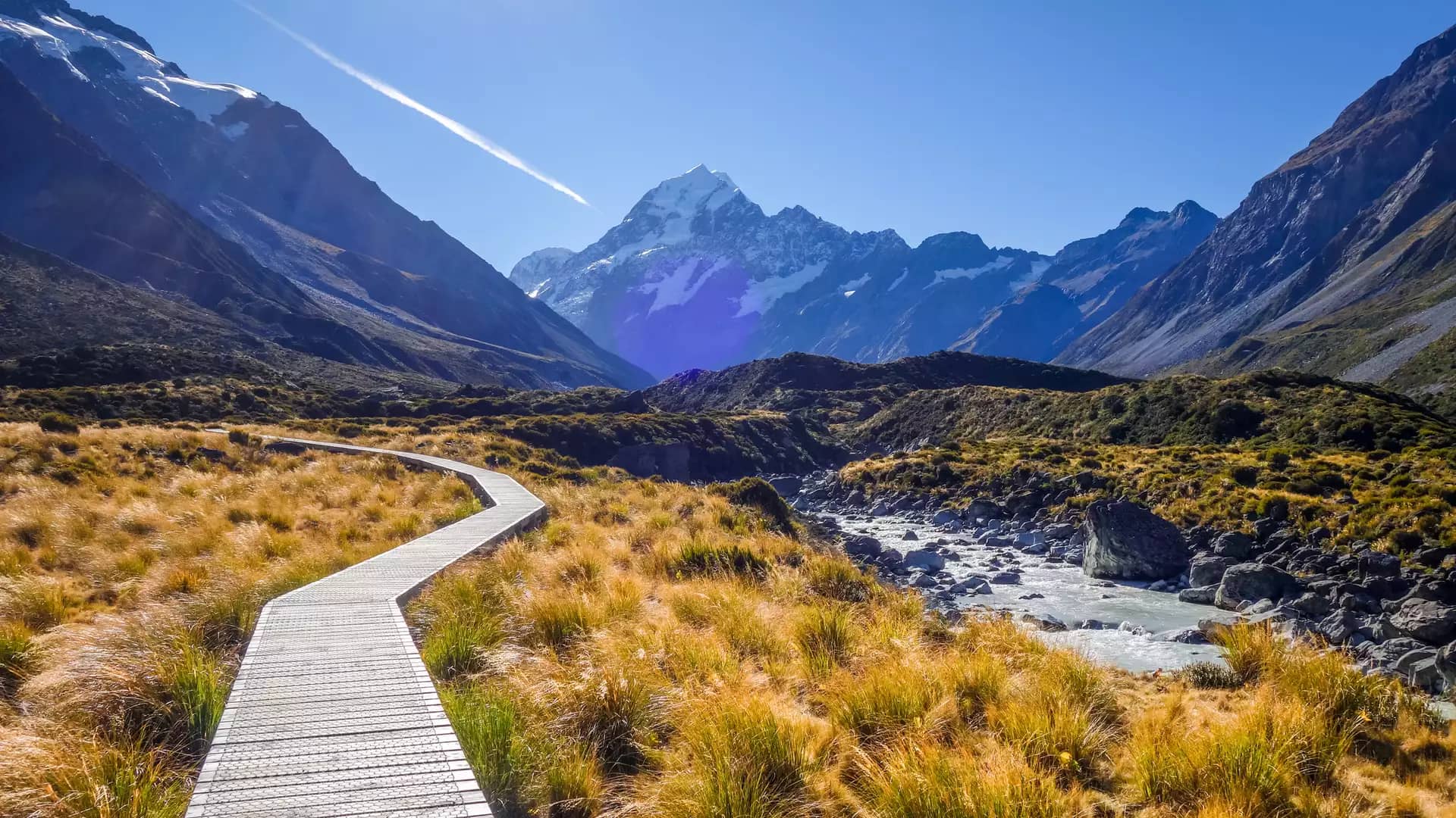 Boardwalk leading through grassland to snow-capped Aoraki Mount Cook under a bright blue sky.