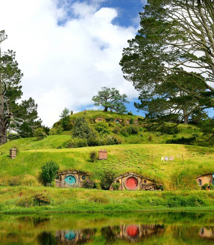Colorful hobbit house doors built into green hills with large trees and a pond reflecting the scene