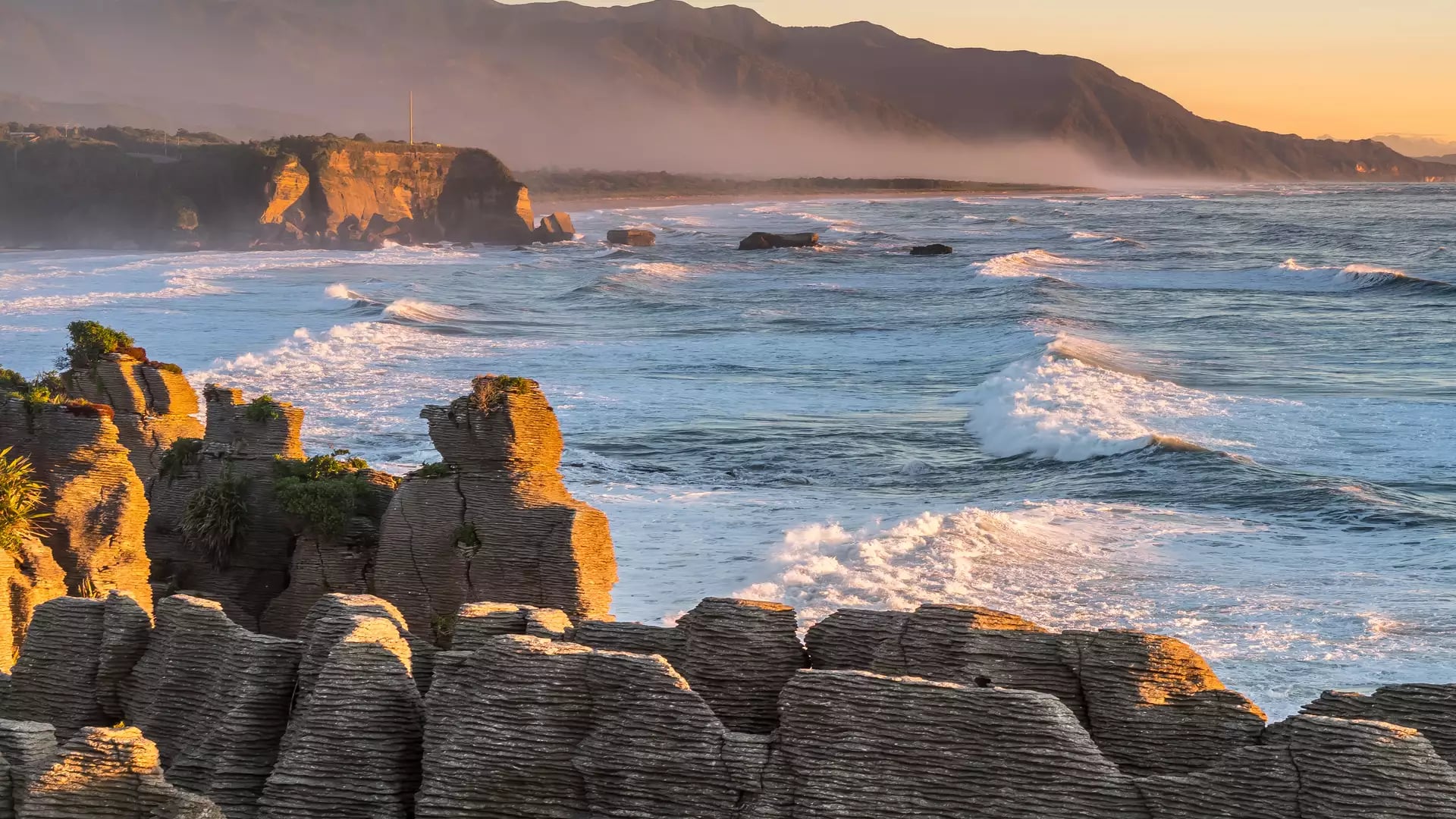 Pancake Rocks with ocean waves and mountains at sunset with mist over the water.