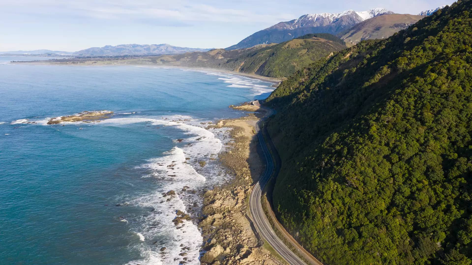 A high-angle aerial view of a winding coastal highway (State Highway 1) sandwiched between steep, forested mountains and a rocky Pacific shoreline under a bright, hazy sun.