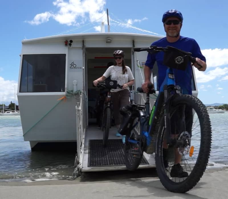 Two cyclists in helmets and sunglasses wheel their bikes down the ramp of the Māpua Ferry onto a sandy beach under a blue sky.