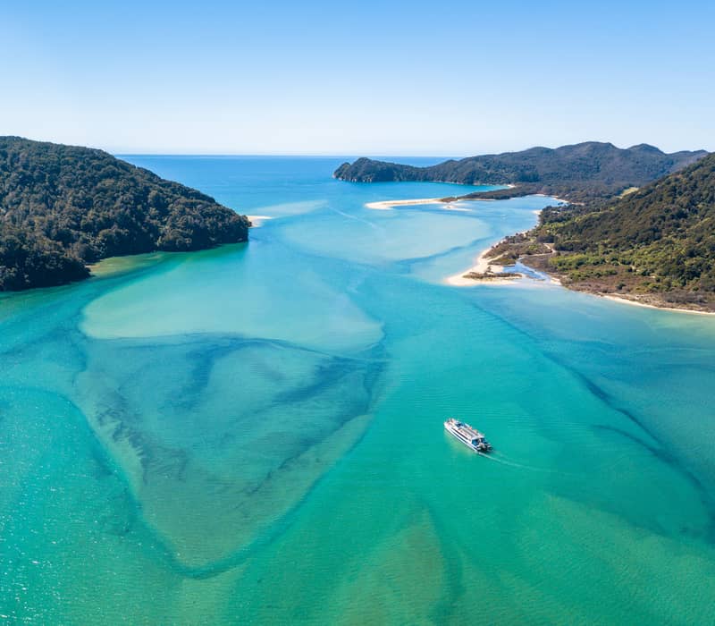 A tour boat sailing through clear turquoise water with forested coastline and sandy beaches.