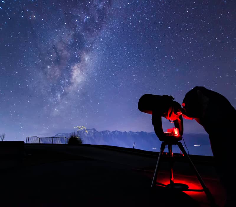 A person looks through a large telescope at a brilliant Milky Way galaxy stretching across a dark purple night sky over the mountains.