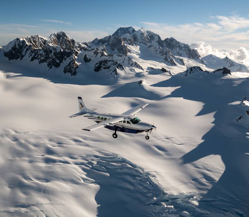 Small plane flying over snowy glacier and rugged mountain peaks under clear sky