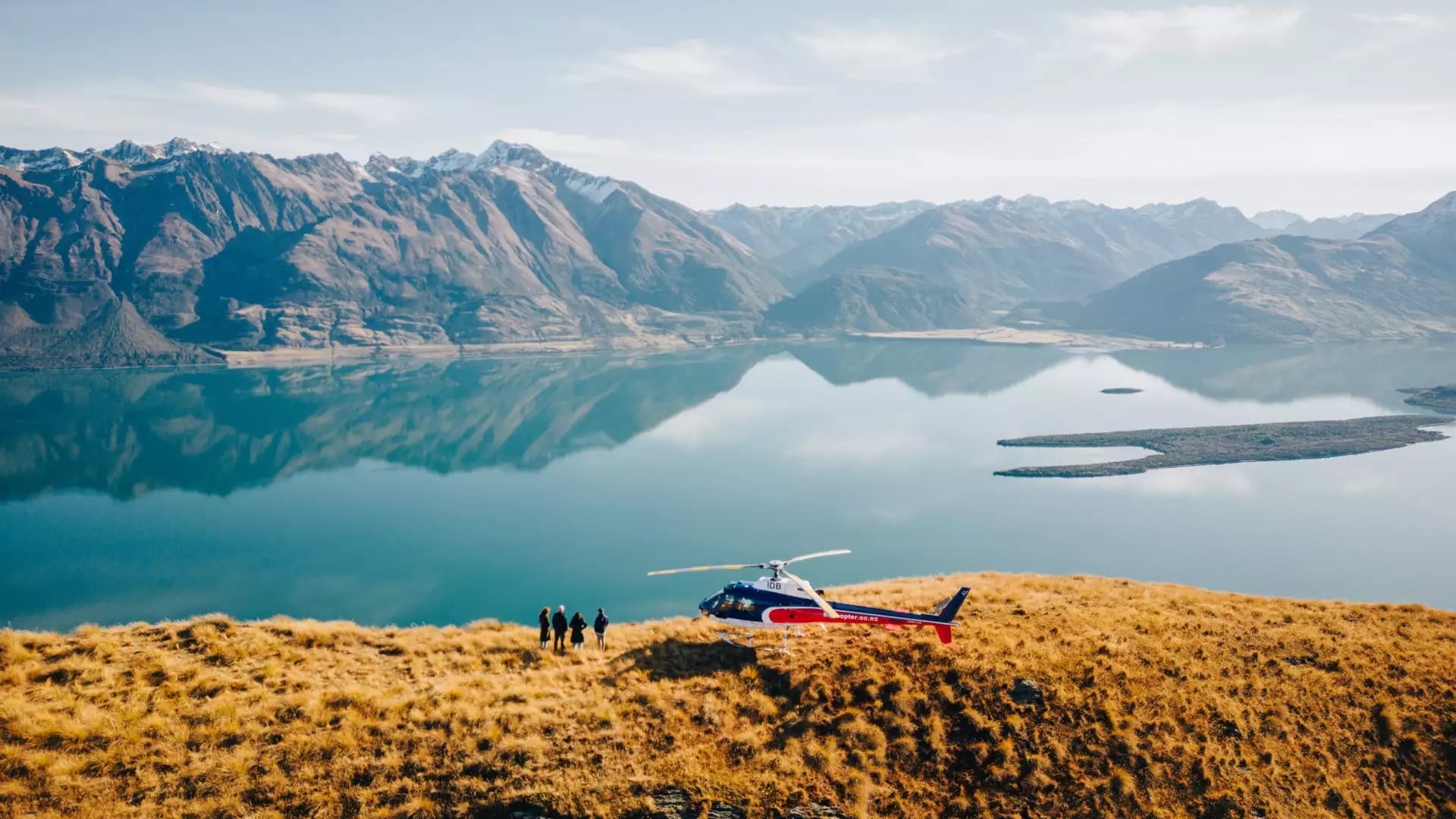 People standing near a helicopter on a hill by a reflective lake and mountains.